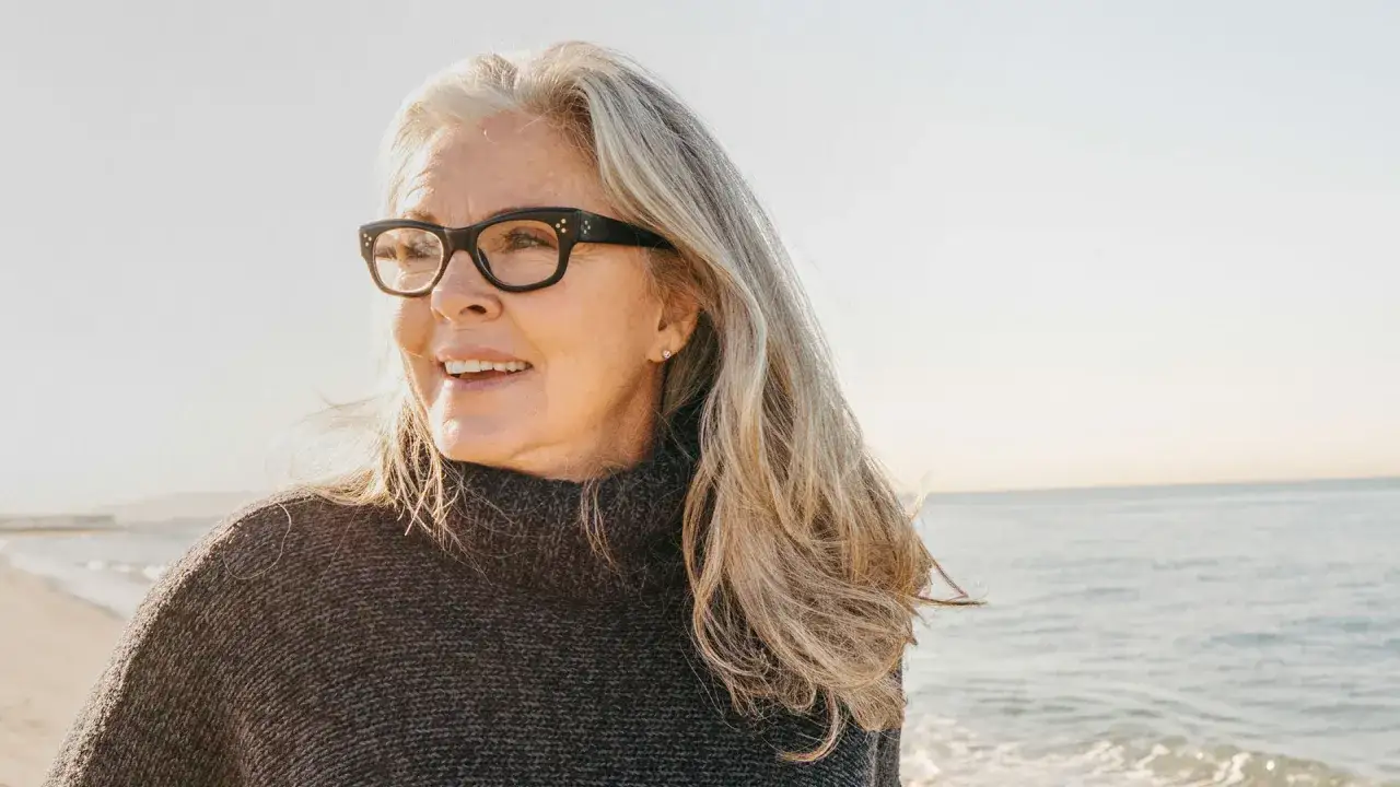 Eine lächelnde Frau mit Brille und langen grauen Haaren am Strand. Ihre Haut zeigt die Spuren des Lebens, sie ist über 60 und strahlt Selbstbewusstsein aus.