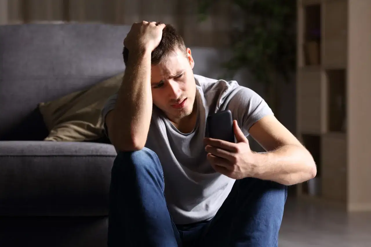 Młody mężczyzna z **nomofobią** siedzi na podłodze, trzymając telefon, z ręką na głowie, wyrażając zmartwienie.