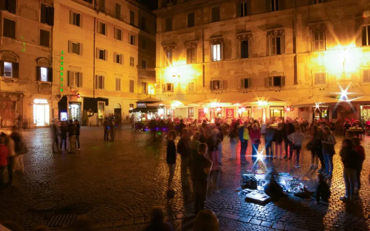 La movida a Roma prende vita in una piazza illuminata, con persone che passeggiano tra locali e ristoranti, creando un'atmosfera vibrante.