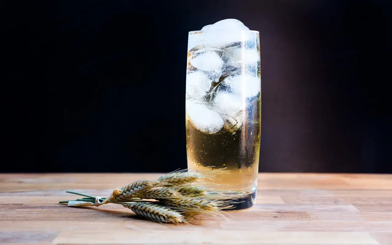 Ein erfrischender Whisky Soda mit Eisw&uuml;rfeln, daneben ein B&uuml;ndel Weizen&auml;hren auf Holztisch.