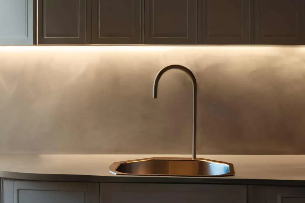 Rubinetto moderno su lavello in metallo, con parete di fondo in **resina dietro cucina** illuminata da una luce led.