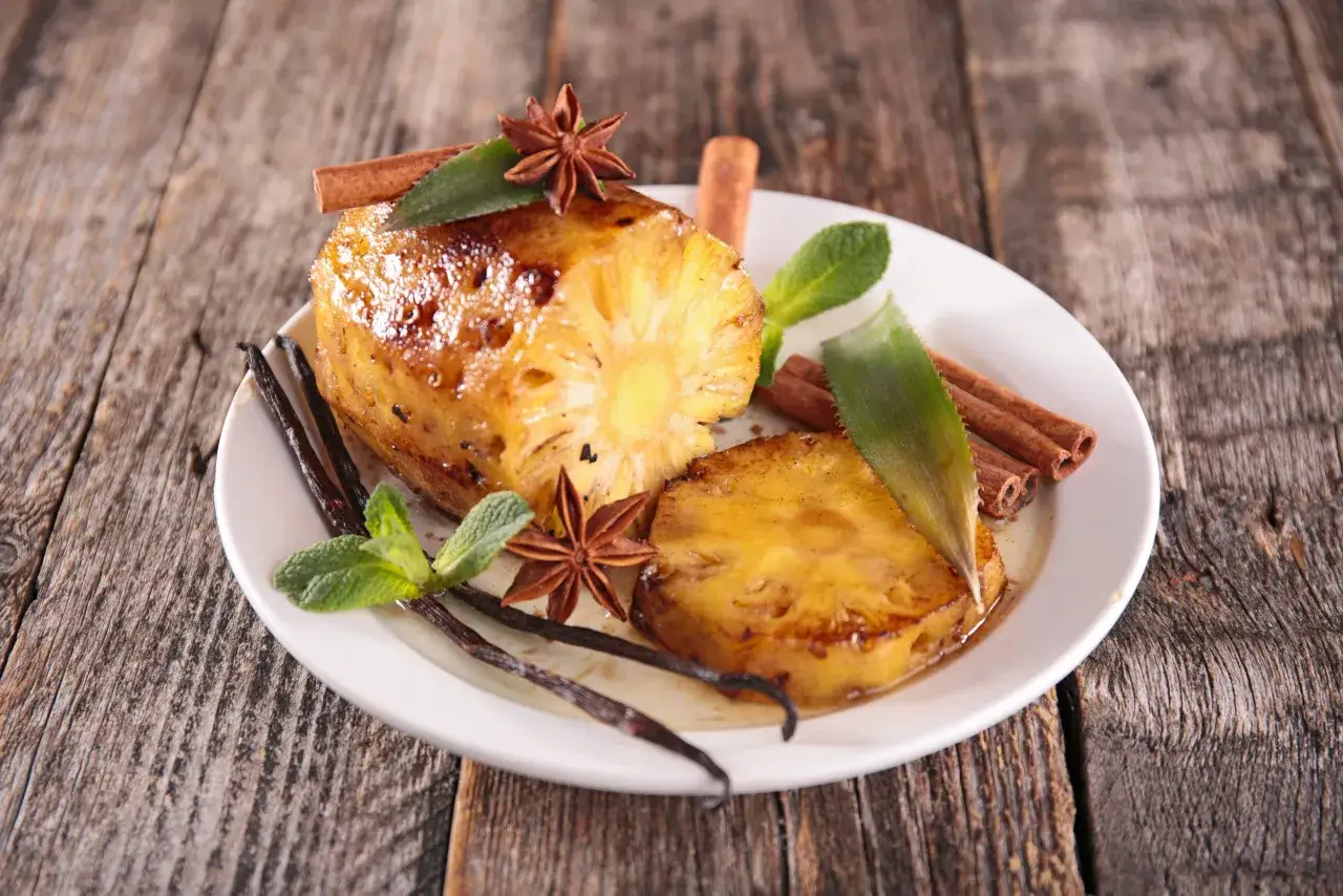 Tranches d'ananas rôti caramélisées, garnies de bâtons de cannelle, d'anis étoilé, de feuilles de menthe et de gousses de vanille sur une assiette blanche.