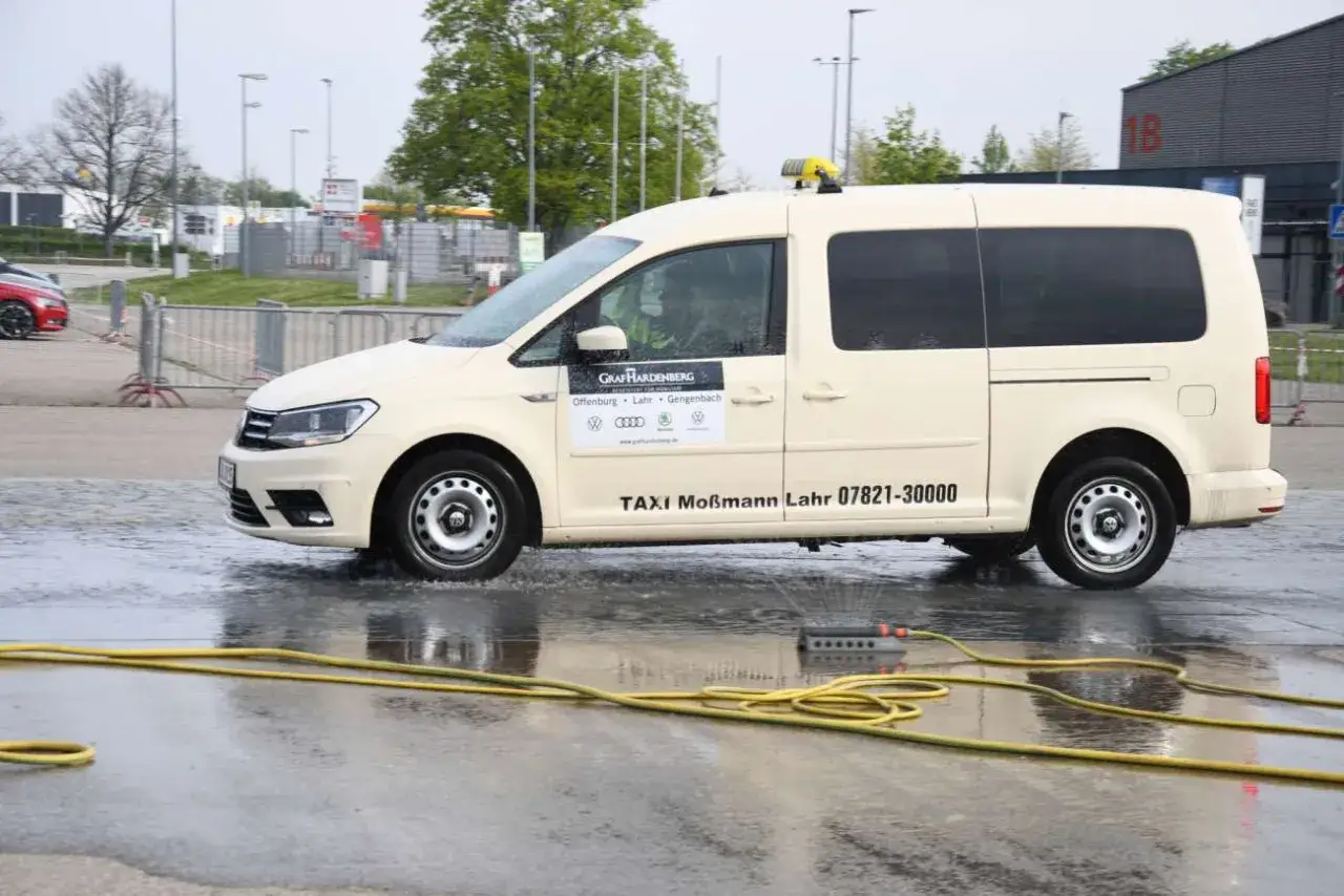Beige Taxi Mo&szlig;mann f&auml;hrt durch Wasser. Ein gelber Schlauch liegt auf dem nassen Boden. Die Berufsgenossenschaft Taxi sorgt f&uuml;r Sicherheit.