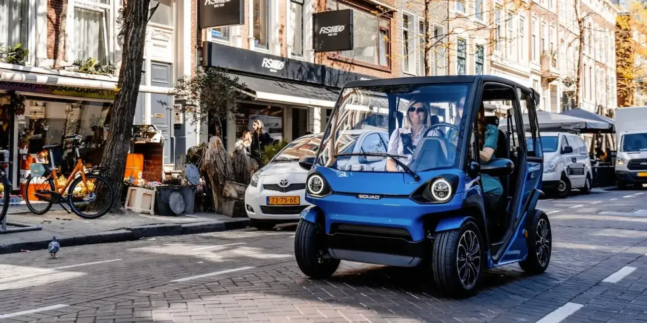 Niebieski, miejski pojazd elektryczny SQUAD, idealny dla szukających najtańszych samochodów elektrycznych, jedzie ulicą.