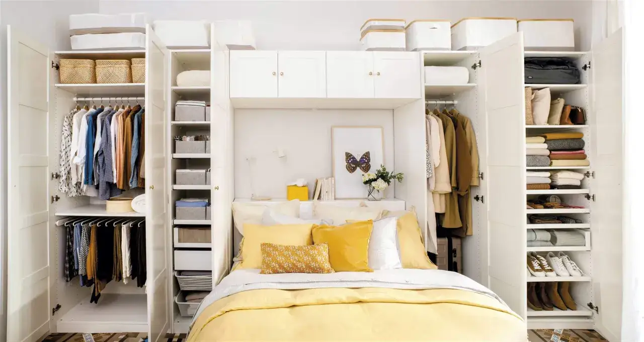 Cama con almohadas amarillas y blancas, rodeada de armarios blancos llenos de ropa y cestas. El espacio entre cama y armario est&aacute; bien aprovechado.