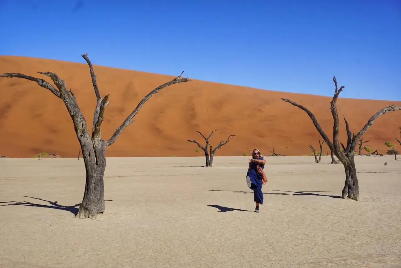 Niesamowita namibia wycieczka! Kobieta w niebieskiej sukience tańczy wśród martwych drzew na tle pomarańczowej wydmy.