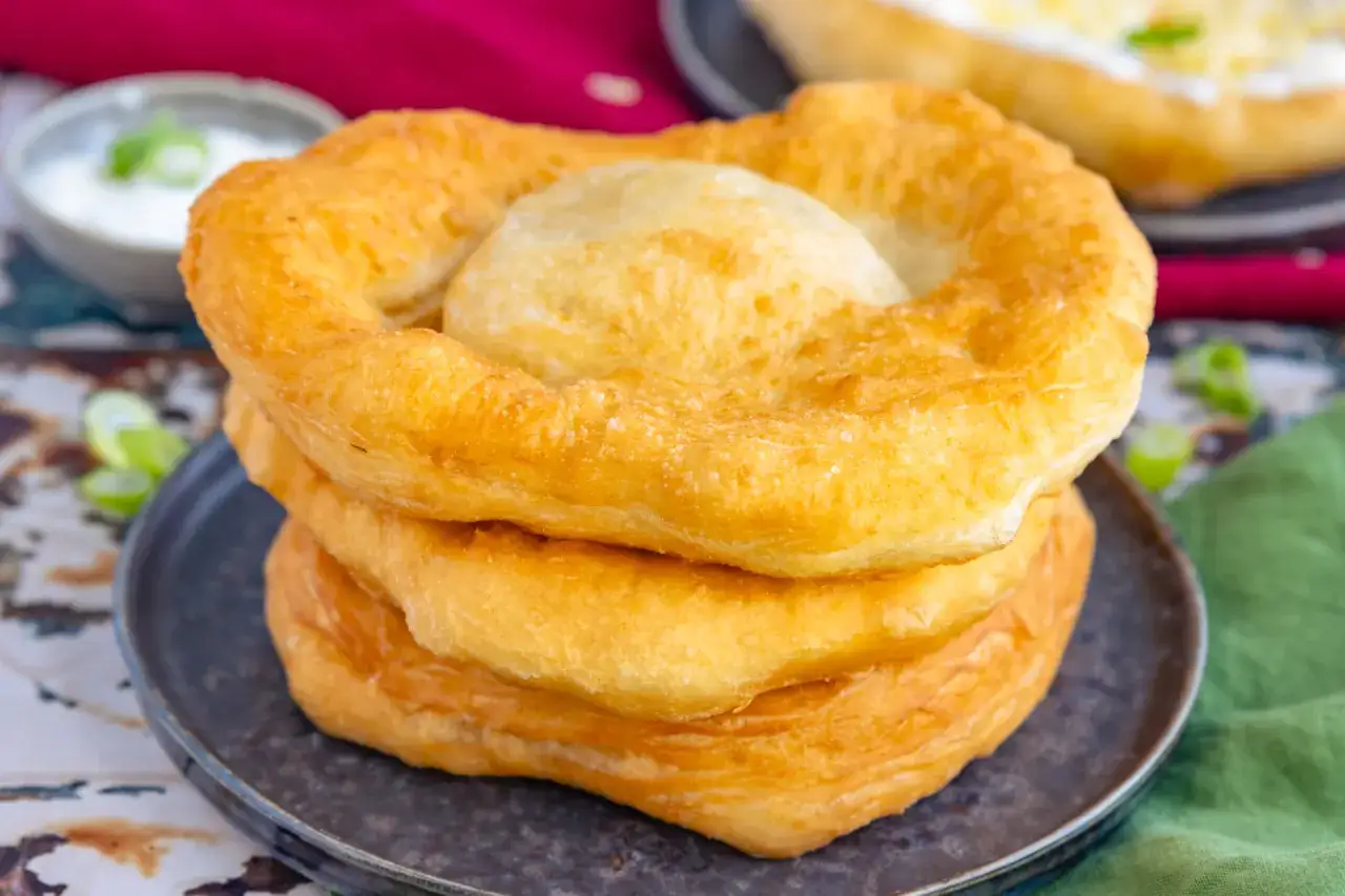 Goldbrauner, frittierter Langos Teig stapelt sich auf einem Teller. Daneben ein Sch&auml;lchen mit Dip und Limettenspalten.