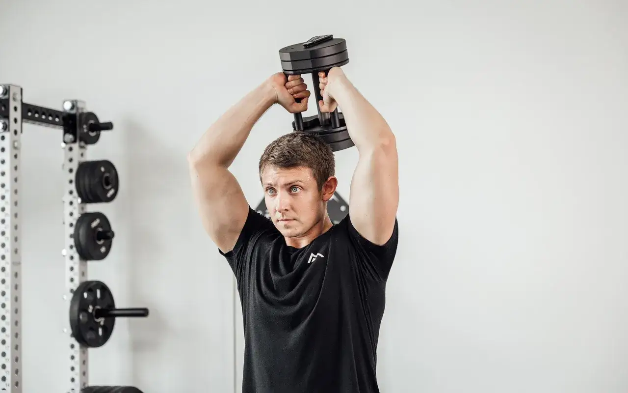 Homme concentr&eacute; effectuant un exercice triceps halt&egrave;re au-dessus de la t&ecirc;te, avec un rack de musculation en arri&egrave;re-plan.