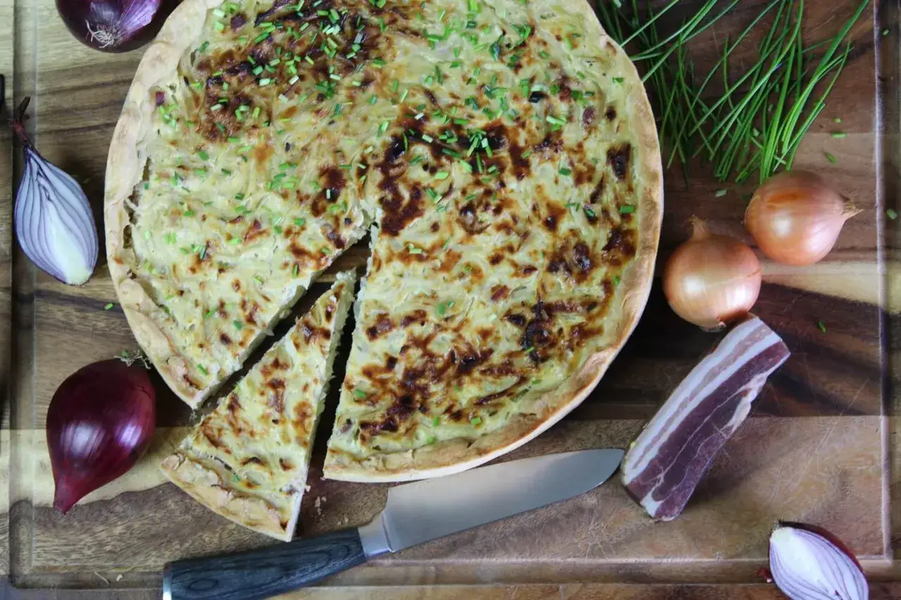 Ein goldbrauner Zwiebelkuchen, frisch aus dem Ofen, mit Schnittlauch bestreut. Daneben liegen Zwiebeln, Speck und ein Messer. Ein echtes zwiebelkuchen originalrezept.