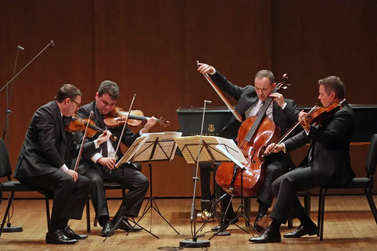 Vier Musiker spielen klassische Instrumente: zwei Violinen, ein Cello und ein Klavier im Hintergrund.