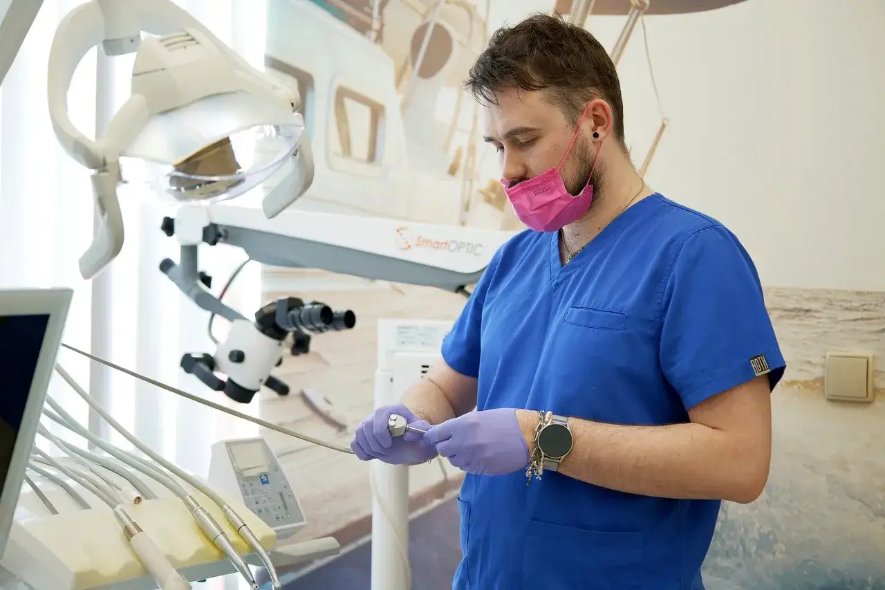 Dentysta w r&oacute;żowej masce przygotowuje narzędzia do zabiegu, być może by zaradzić obluzowaniu korony zęba.