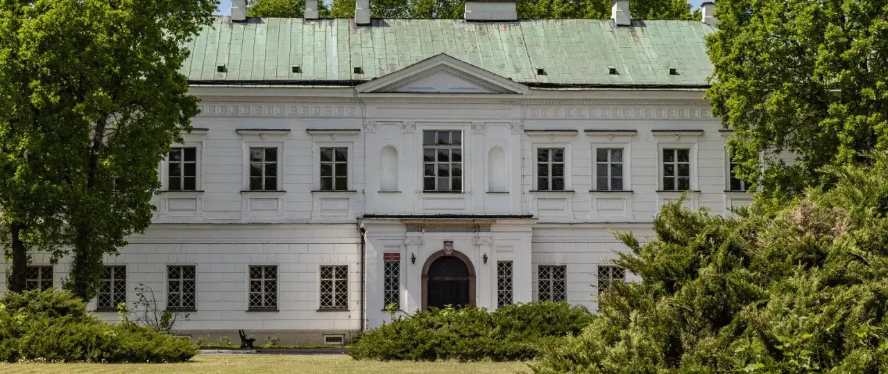 Biały pałac w Falentach z zielonym dachem, otoczony bujną zielenią drzew i krzewów.