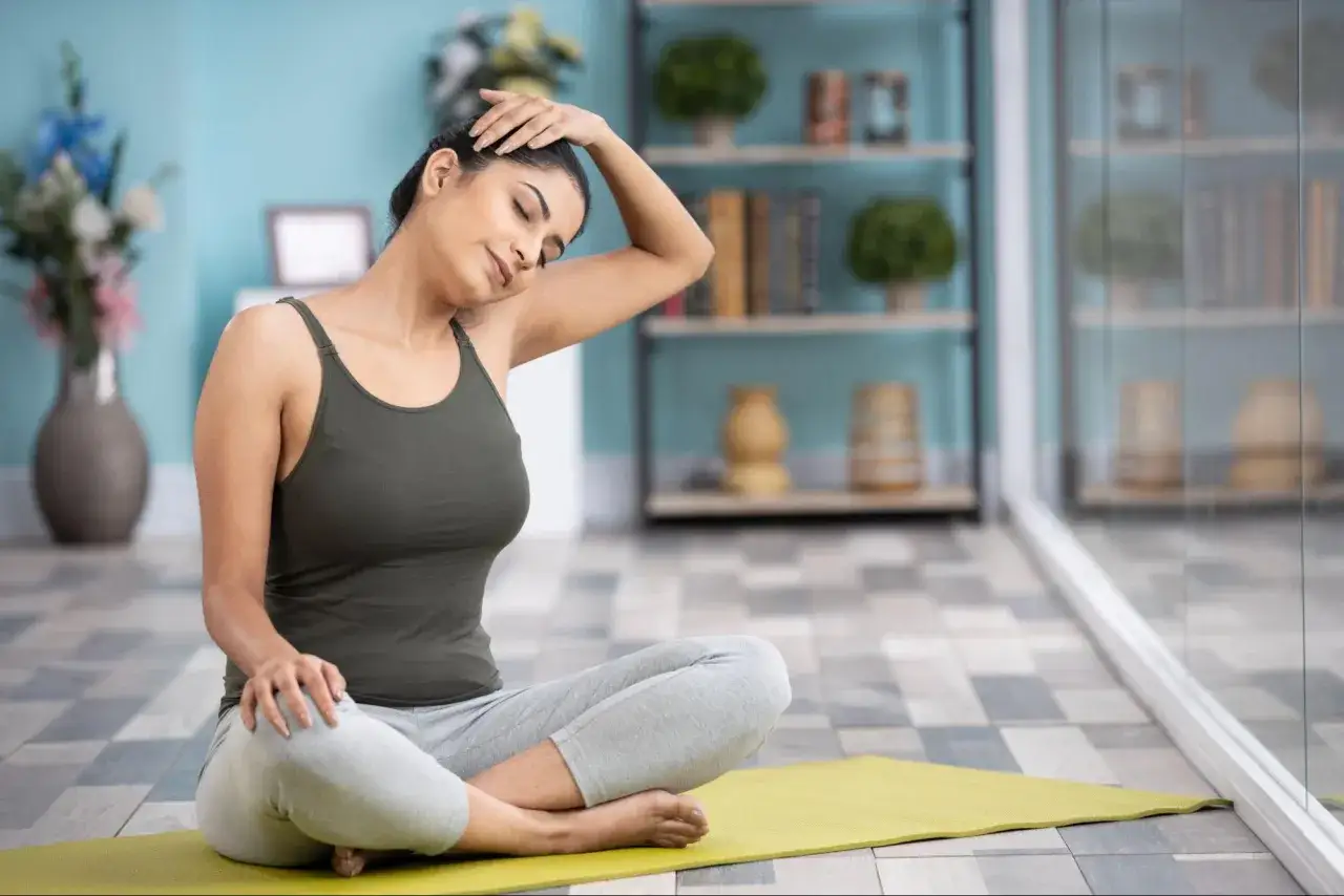 En kvinna sitter på en yogamatta och sträcker försiktigt sin nacke för att slappna av i nacken.