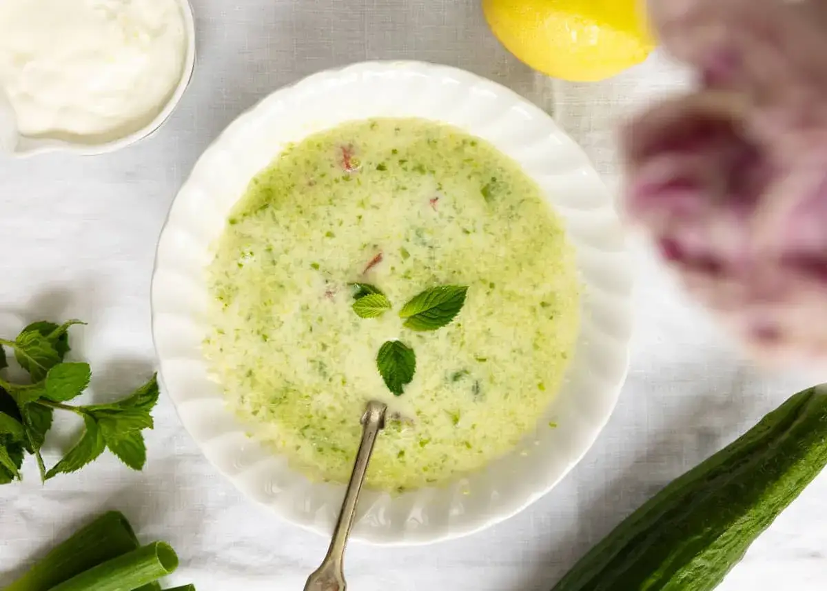 Une assiette de soupe de concombre cr&eacute;meuse, garnie de menthe fra&icirc;che, avec des ingr&eacute;dients frais comme un concombre et un citron &agrave; proximit&eacute;.