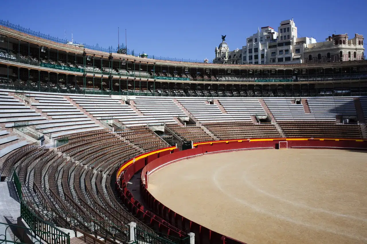 Vista interior de una plaza de toros vac&iacute;a, con gradas de madera y hormig&oacute;n, arena de tierra y edificios urbanos al fondo. &iexcl;Una visita a la plaza de toros!