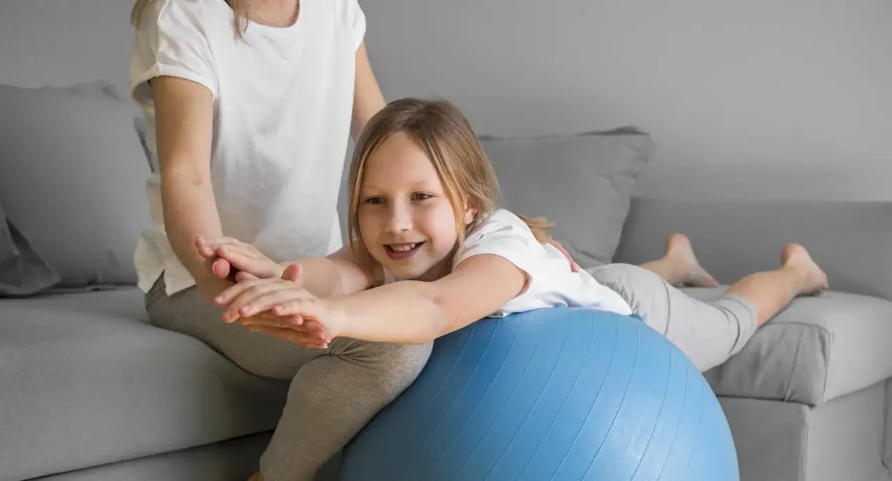 Dziewczynka z obniżonym napięciem mięśniowym ćwiczy mowę na piłce rehabilitacyjnej z pomocą terapeuty.