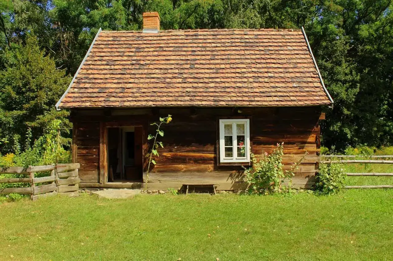 Drewniana chata z glinianym dachem, typowa dla skansenów w Polsce, otoczona zielenią i drewnianym płotem.