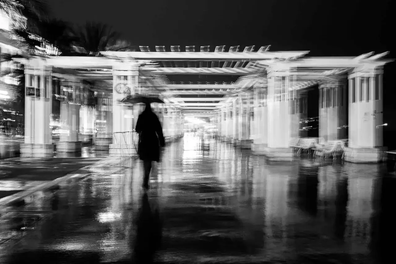 Une silhouette solitaire sous un parapluie traverse une all&eacute;e de **photo de rue** mouill&eacute;e et r&eacute;fl&eacute;chissante, sous une structure architecturale floue.
