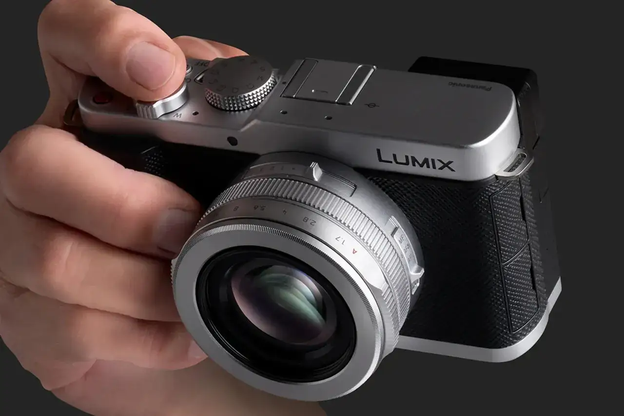 En hand h&aring;ller en silverf&auml;rgad Panasonic LX100-kamera med en objektivring som visar bl&auml;ndartal.