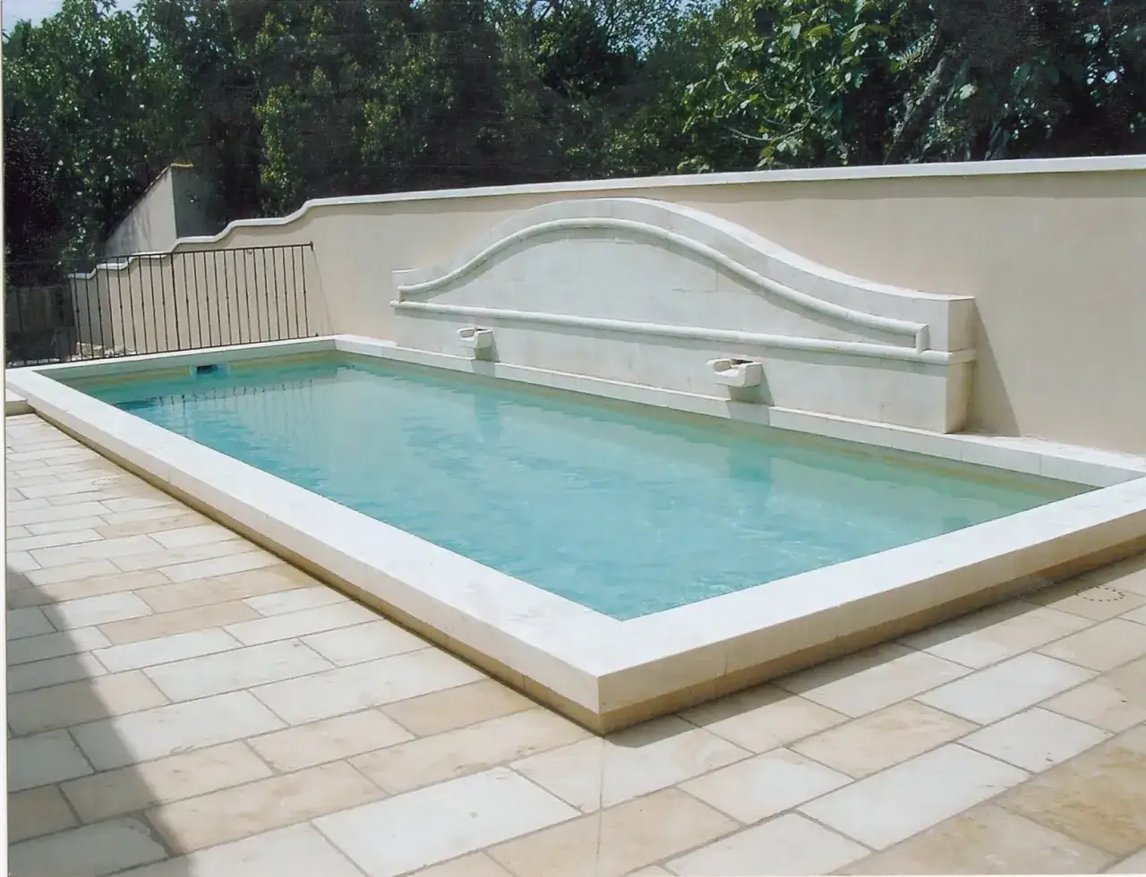 Une piscine rectangulaire aux marges en pierre, entourée d'un dallage clair. Un mur orné d'une fontaine en pierre domine le bassin.