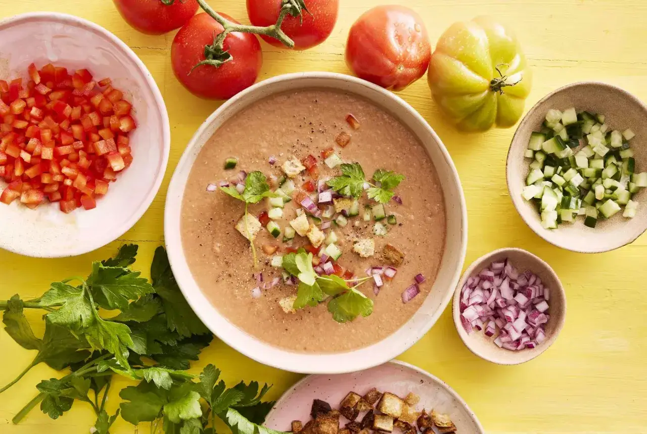 En skål med gazpacho, toppad med krutonger, gurka och lök. Ingredienser för gazpacho recept syns runt om.