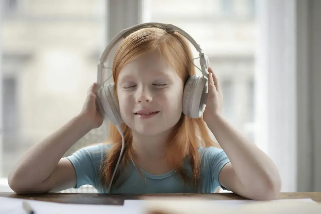 Dziewczynka z rudymi włosami słucha fajnych audiobook&oacute;w dla młodzieży przez białe słuchawki, z zamkniętymi oczami i uśmiechem.