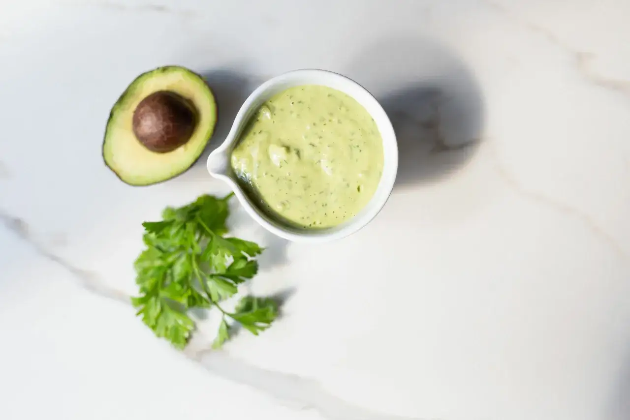 Eine Schale mit cremiger Avocado-Creme, daneben eine halbe Avocado und Petersilie auf Marmor.