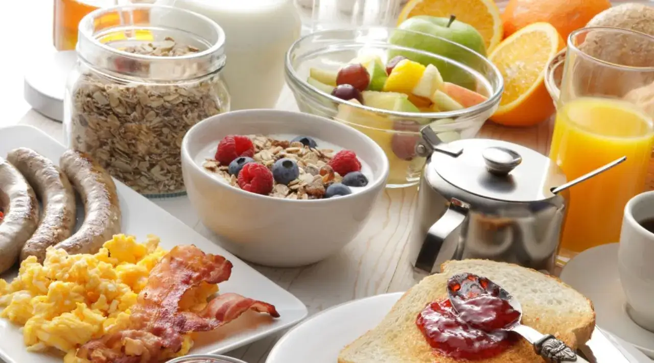 Un petit déjeuner prise de masse copieux : œufs brouillés, bacon, saucisses, yaourt aux fruits, muesli, jus d'orange et toasts à la confiture.