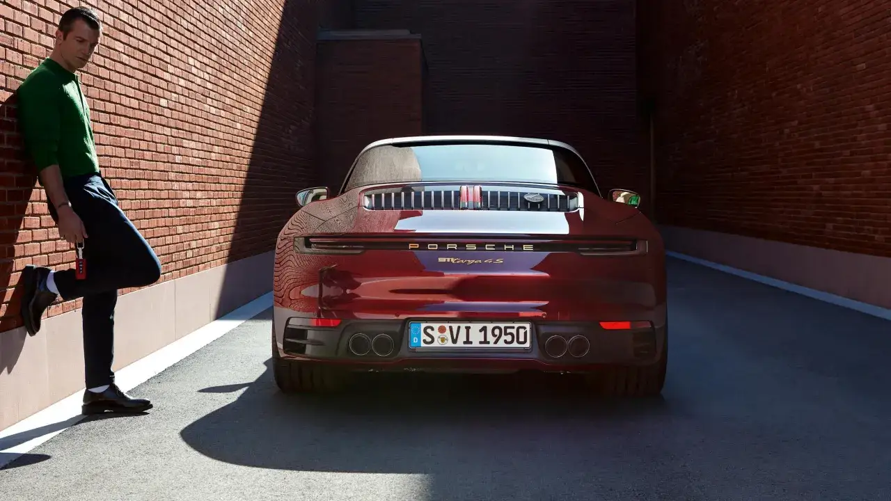 Ein Mann lehnt an einer Ziegelwand neben einem roten Porsche von hinten.
