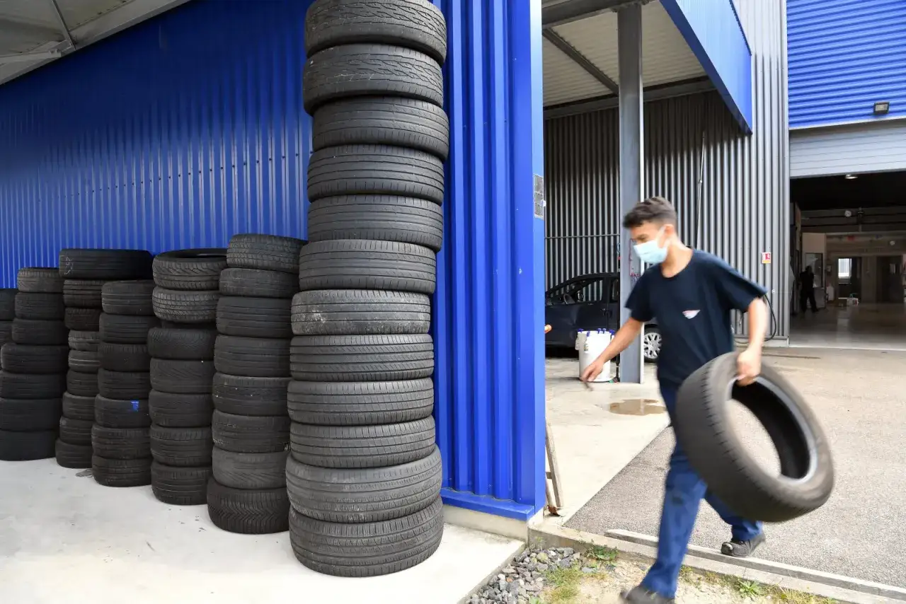 Un jeune homme masqu&eacute; transporte un pneu usag&eacute;, pr&ecirc;t &agrave; le jeter. Des piles de pneus s'alignent le long d'un mur bleu.