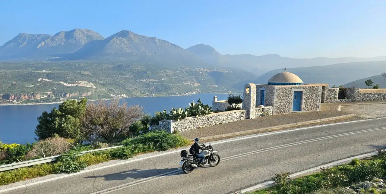 Motocyklista przemierza malowniczą grecką drogę z widokiem na zatokę i góry. Czy Grecja jest droga? Podróżowanie po niej może być przystępne, jeśli wybierze się odpowiednie środki transportu.