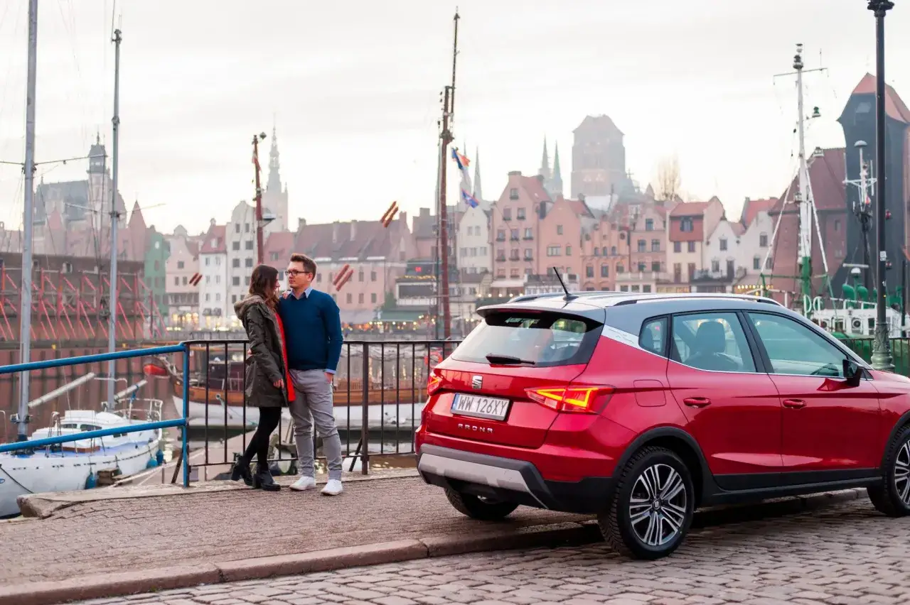 Para spaceruje obok czerwonego Seat Arona 1.0 TSI, z widokiem na zabytkowe kamienice i żaglowce.