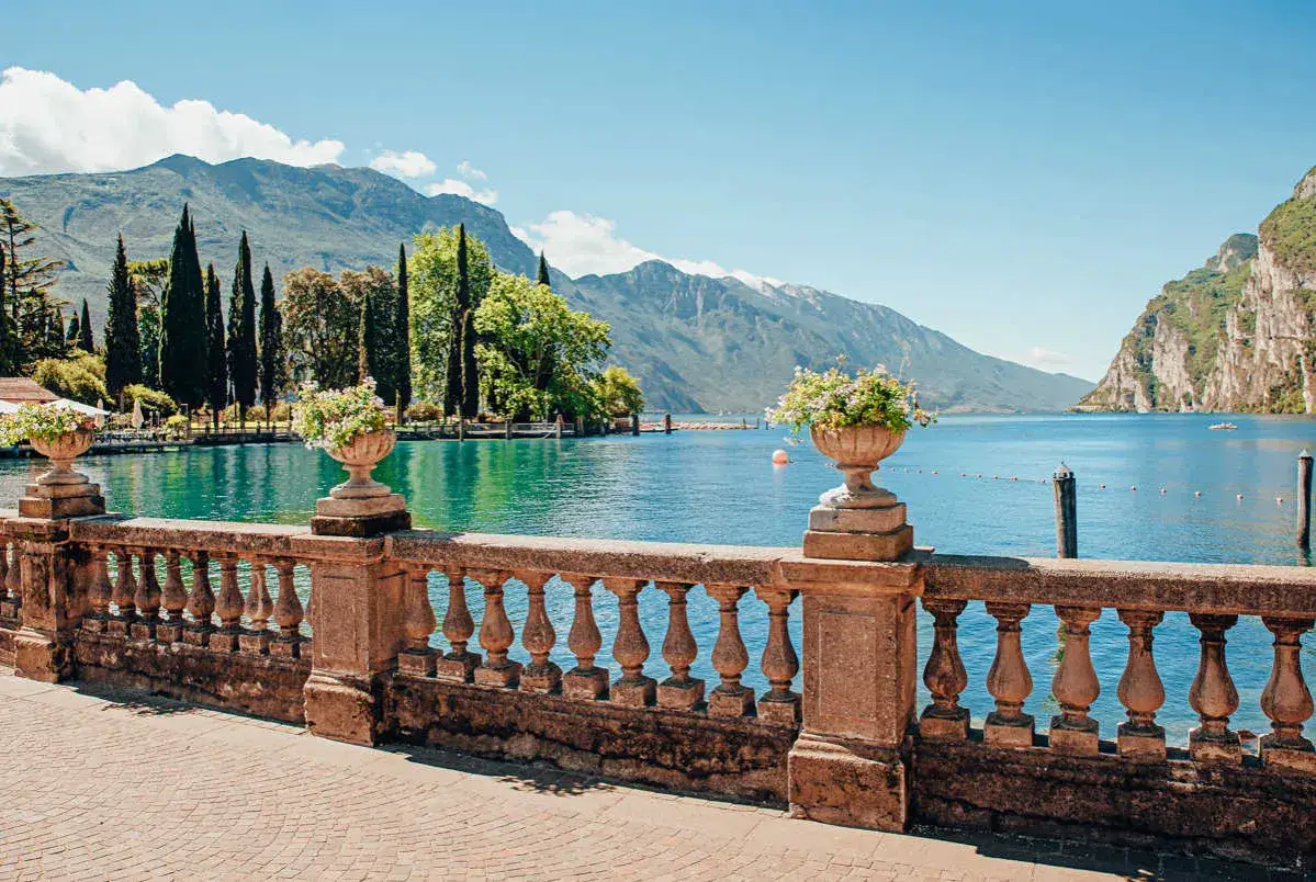 Kamienne balustrady i wazony z kwiatami nad turkusowym jeziorem Garda, otoczonym zielonymi wzgórzami i górami.