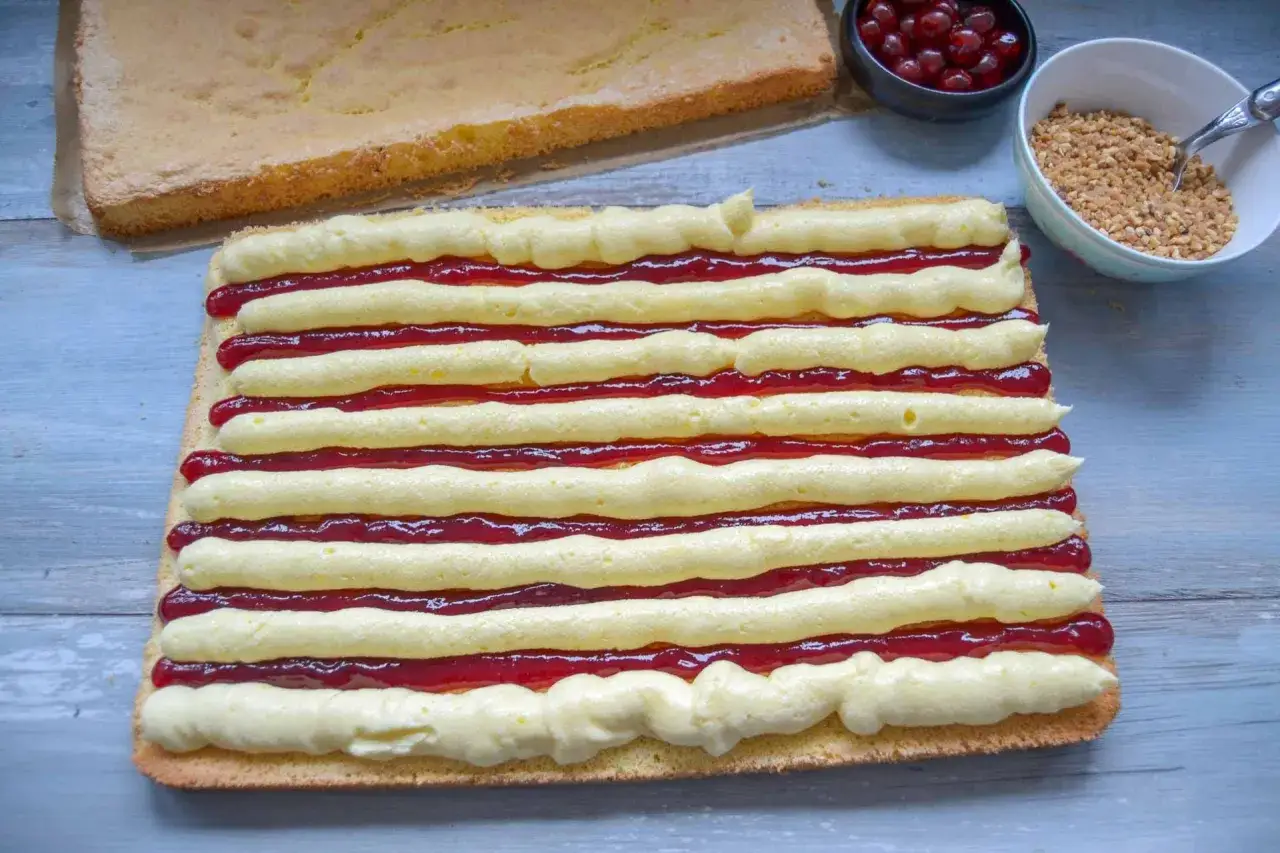 Ein Frankfurter Kranz Blech wird mit Buttercreme und Marmelade belegt. Daneben liegen Kirschen und gehackte Nüsse.