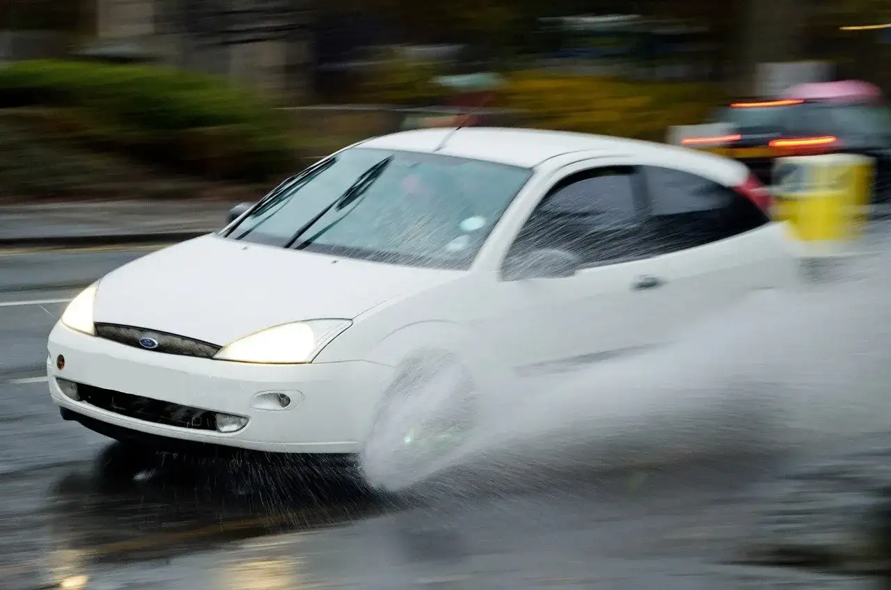 Auto bianca in movimento su strada bagnata, con le ruote che sollevano un'enorme quantità d'acqua. Questo è un esempio di aquaplaning, cos'è: perdita di aderenza dovuta all'acqua.