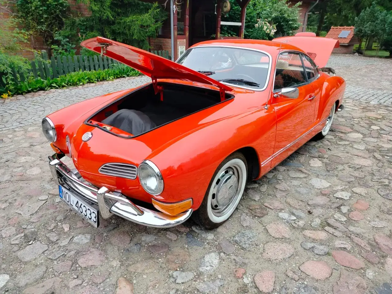 Pomarańczowy Karmann Ghia z otwartą maską i bagażnikiem, lśniącym na brukowanym podwórku.