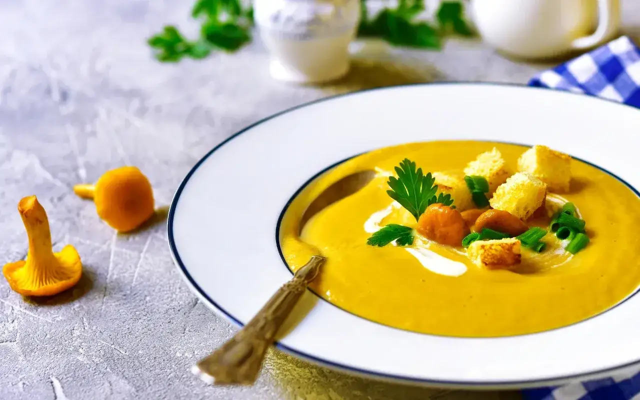 Aksamitna zupa krem z kurek, udekorowana grzankami, natką pietruszki i szczypiorkiem. Obok leżą świeże kurki.
