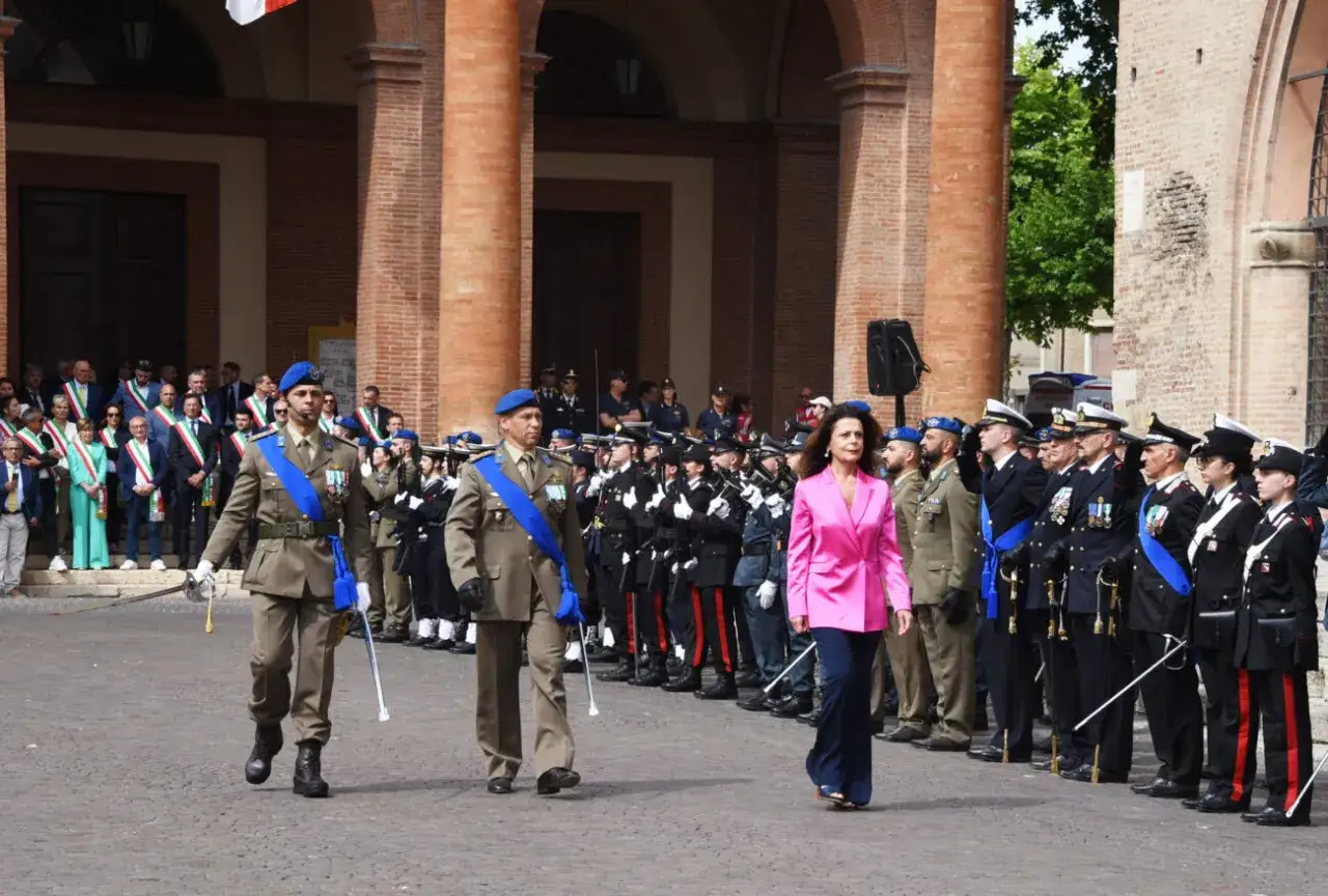 Parata militare a Rimini il 2 giugno. Uomini in divisa e una donna in giacca rosa camminano tra le colonne di un edificio storico.