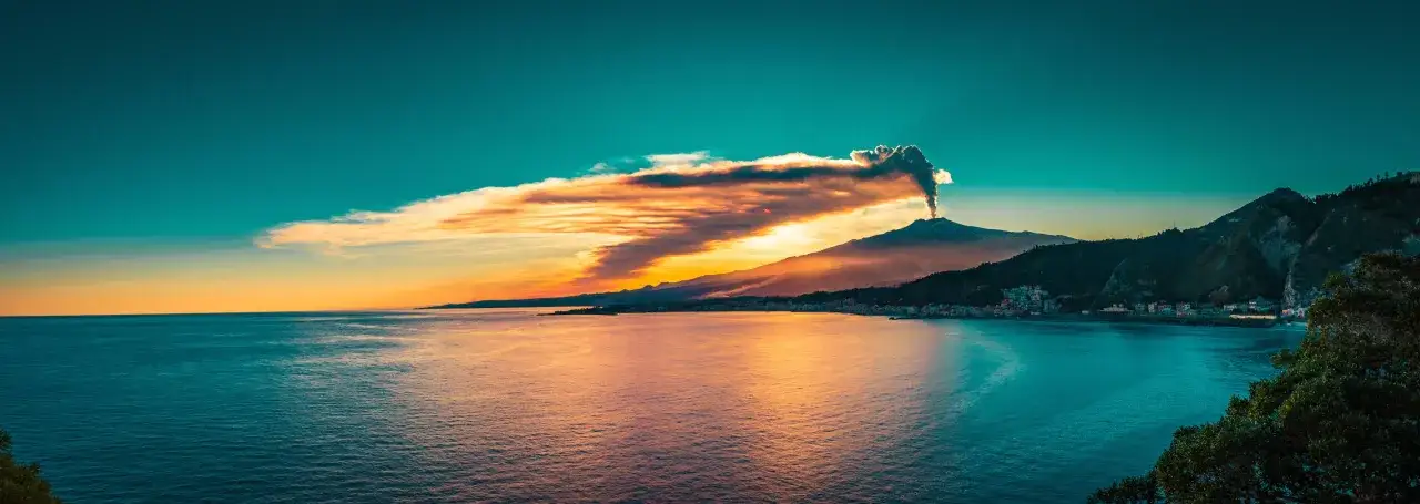Etna fumante al tramonto, un'immagine iconica dei luoghi più belli della Sicilia. Il mare riflette i colori caldi del cielo.