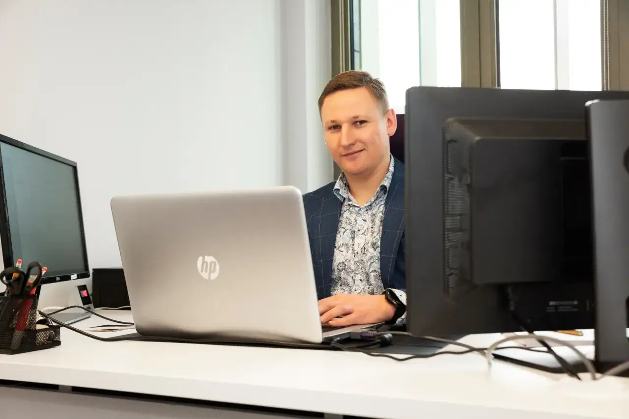 Mężczyzna w garniturze siedzi przy biurku z laptopem i monitorami.