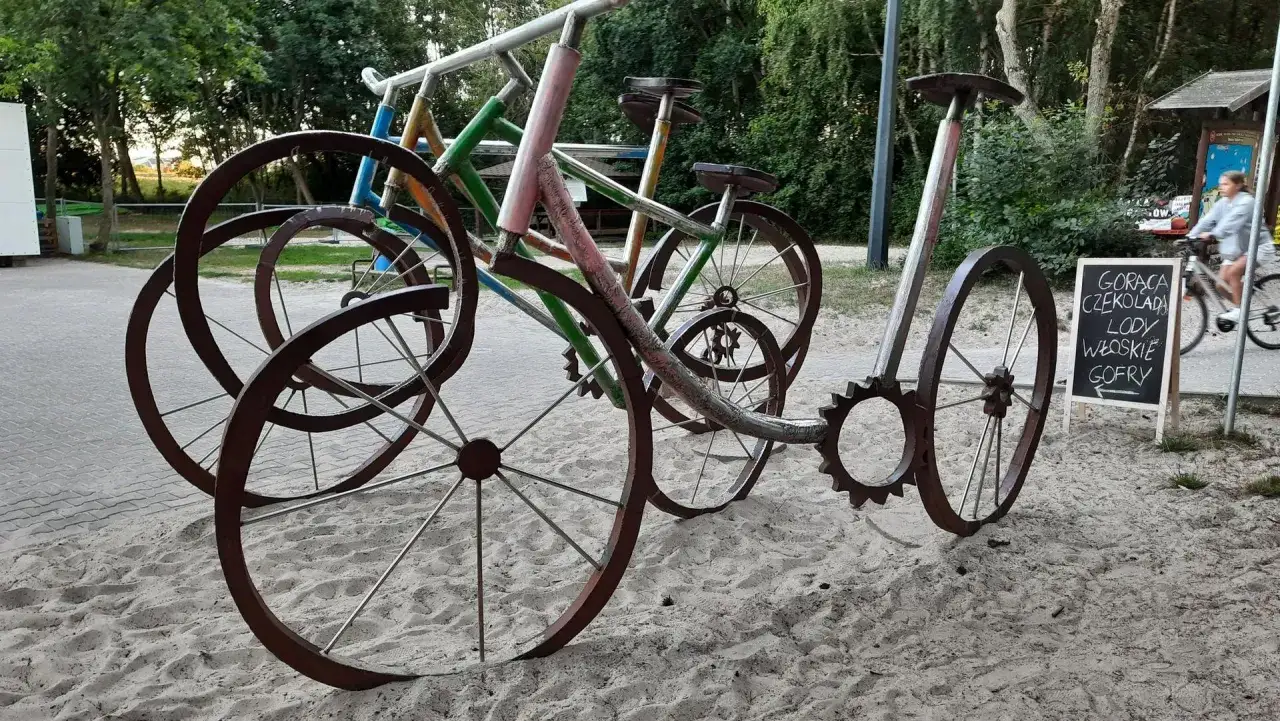 Metalowe rowery-rzeźby na piasku, zapraszają do skorzystania z atrakcji Grzybowo. Tablica głosi: 