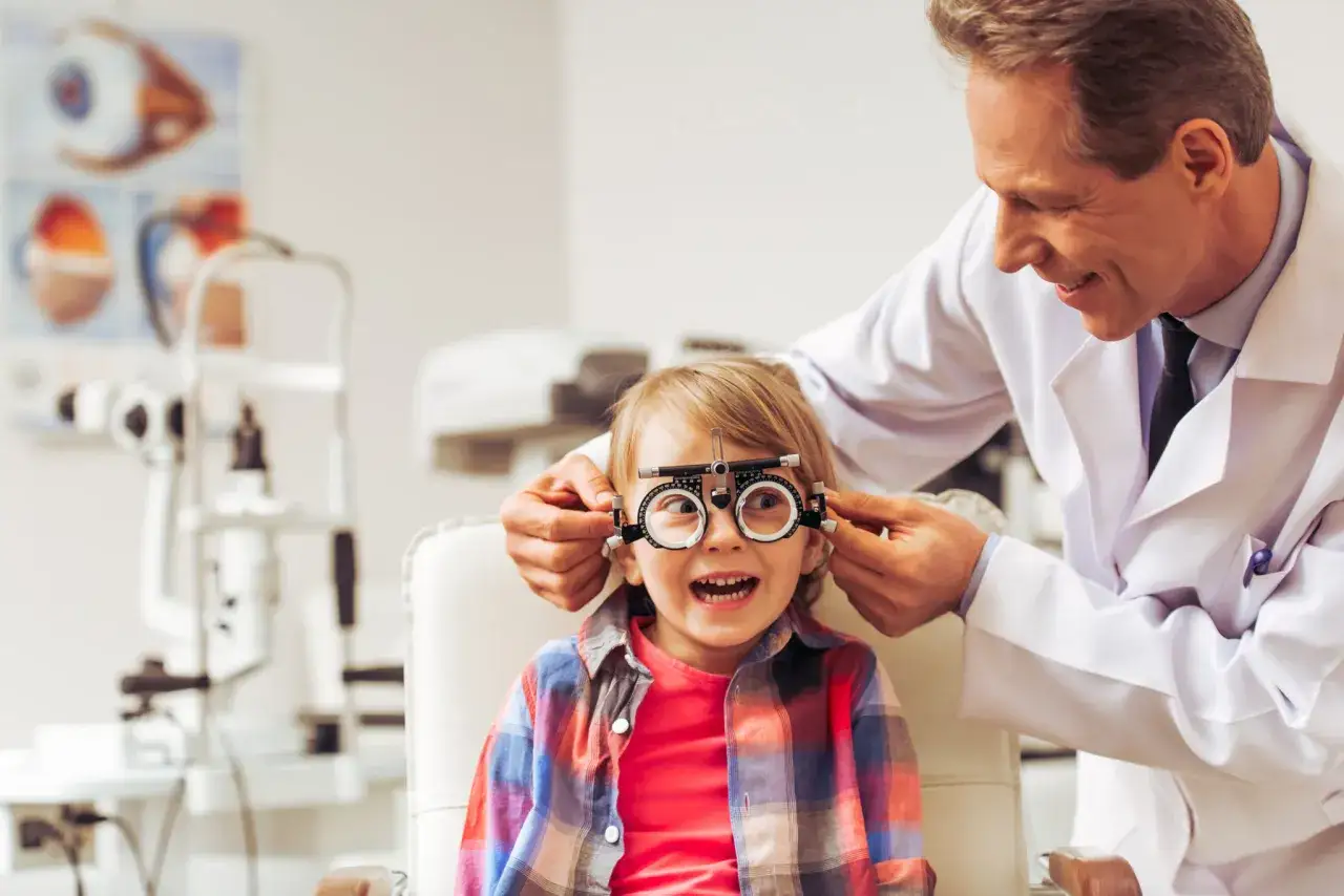 Uśmiechnięty optometrysta przeprowadza badanie wzroku u dziecka. Mały pacjent ma na nosie oprawki próbne, a jego oczy są szeroko otwarte.