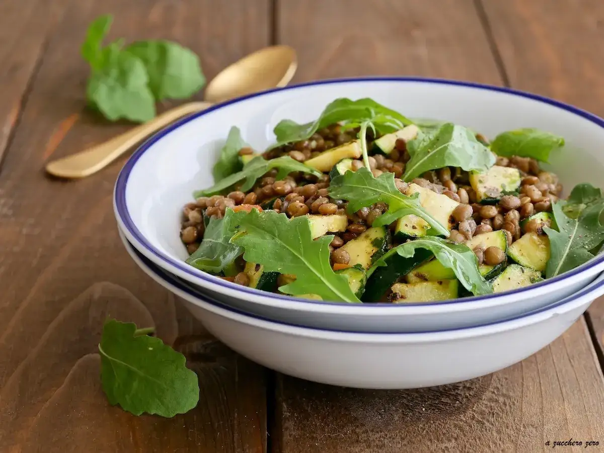 Deliziosa insalata di lenticchie con zucchine e rucola fresca, servita in una ciotola bianca su un tavolo di legno.