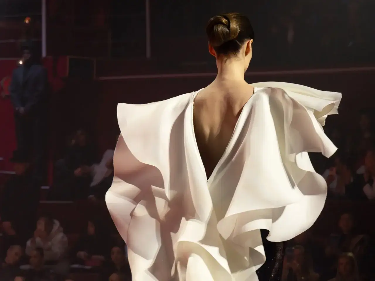 A model showcases a dramatic white fashion couture gown with voluminous ruffles cascading down her back, a vision of elegance on the runway.