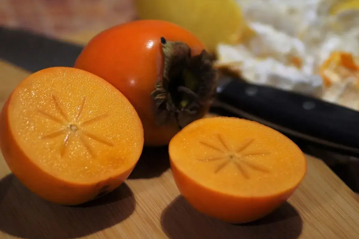 Pomarańczowe owoce kaki, jeden cały, dwa przekrojone, ukazują swoje wnętrze. Warto poznać owoc kaki właściwości.