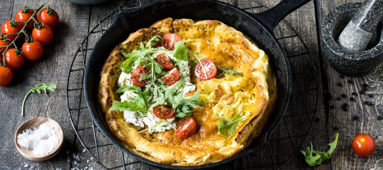Ein goldbrauner Dutch Baby mit Rucola, Kirschtomaten und Ziegenkäse in einer Gusseisenpfanne.