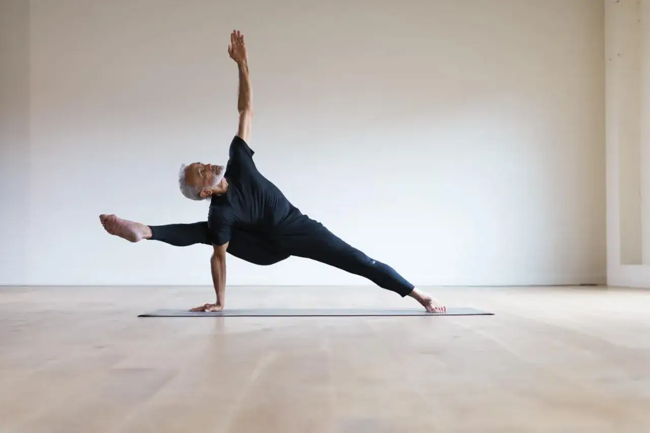 Maestro Dharma Mittra en una postura de yoga desafiante, con una pierna extendida y la otra flexionada, equilibrando su cuerpo sobre una mano.