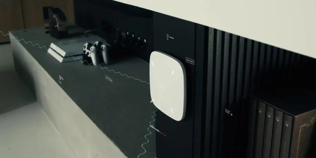 Un appareil blanc, peut-être une alarme anti brouillage, est fixé sur un meuble noir, à côté d'une console de jeux et de livres.