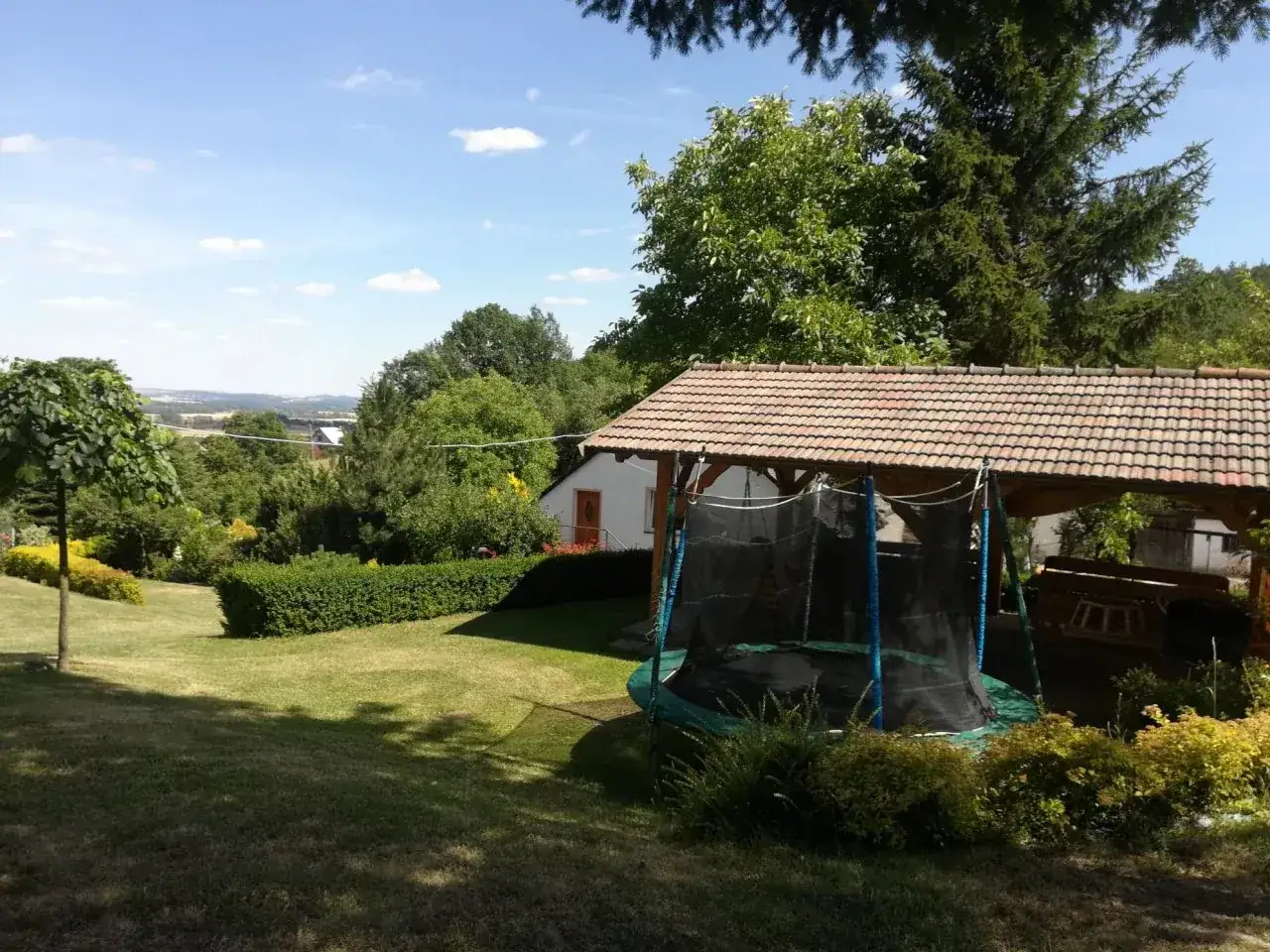 Letnia, słoneczna sceneria w Brzeźnicy. Ogród z trampoliną pod zadaszeniem, zielonymi krzewami i drzewami, w tle rozciąga się malowniczy krajobraz.