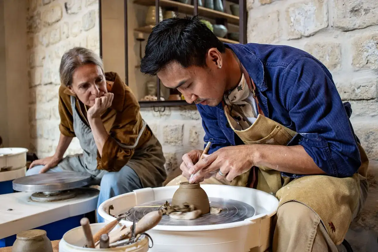 Un artisan qui travaille avec l'argile façonne une poterie sur un tour. Une femme observe attentivement, son menton appuyé sur sa main.